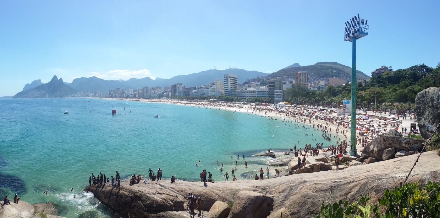A busy Arpoador beach and turquoise water seen from the rocks, with Ipanema and Morro Dois Irmãos in the distance
