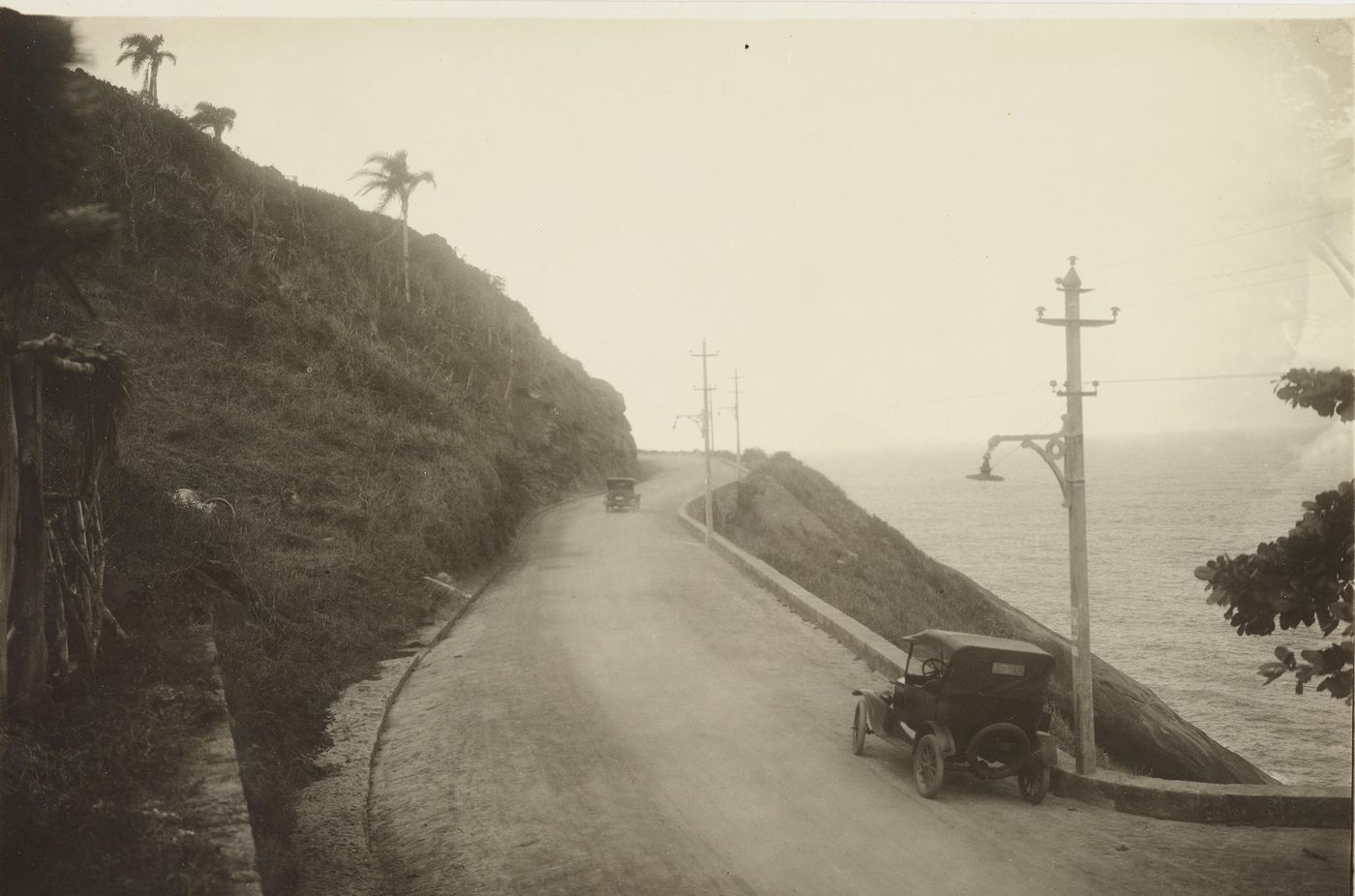 Black-and-white photograph of an early car on Avenida Niemeyer, a narrow cliff road above the sea lined with lamp posts
