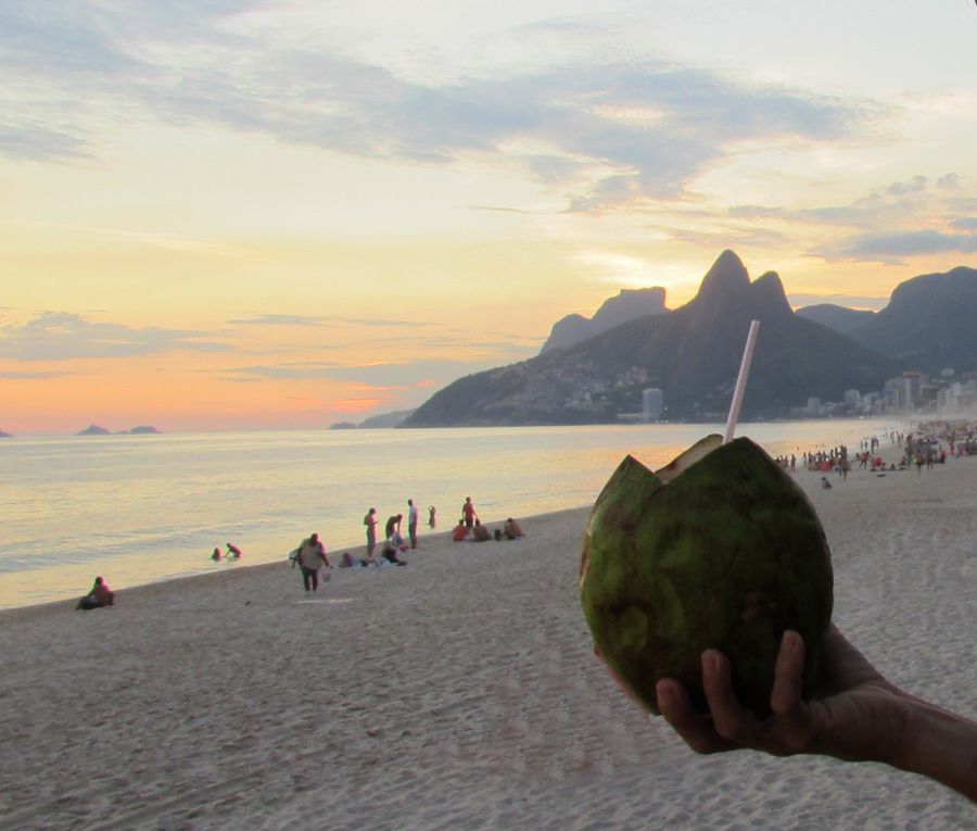A green coconut with a straw held up on Ipanema Beach at sunset, with Morro Dois Irmãos behind