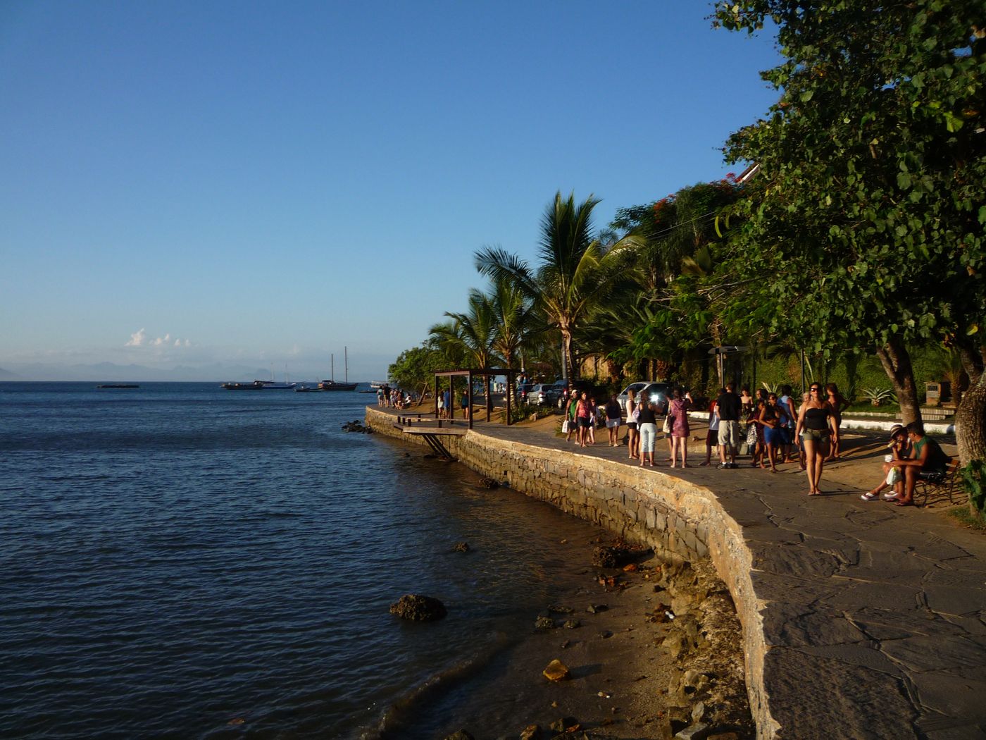 The stone seawall promenade of Orla Bardot in Búzios in late-afternoon light, with people strolling and boats on the bay