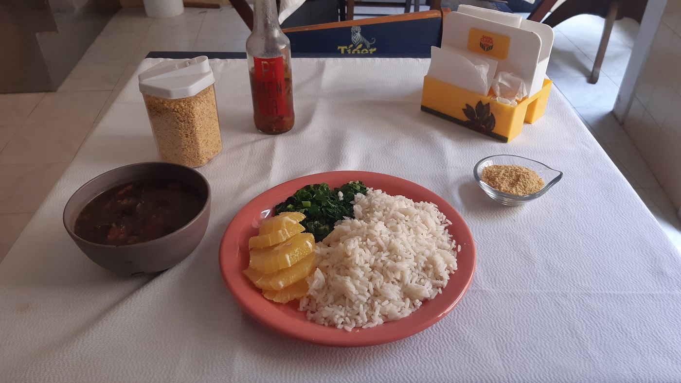 A feijoada lunch on a white tablecloth: the bean stew in a bowl beside a plate of rice, collard greens and orange slices, with farofa on the side