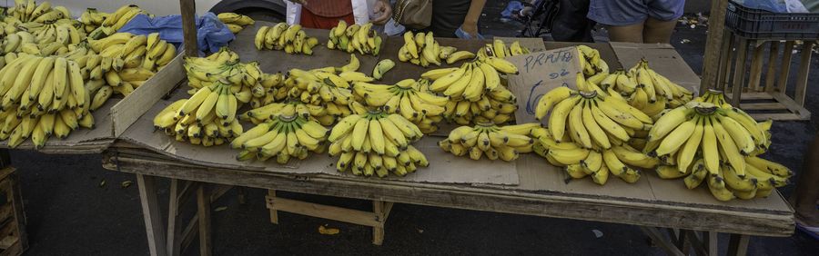 Bunches of bananas piled on a market stall, with the stallholder and shoppers under striped awnings