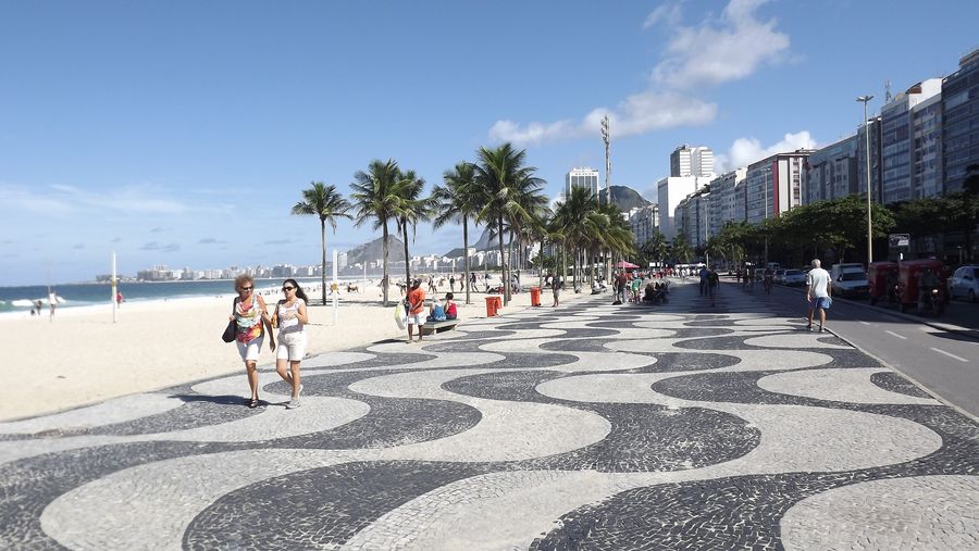 O mosaico de ondas preto e branco do calçadão de Copacabana, com palmeiras, prédios e pessoas caminhando junto à praia