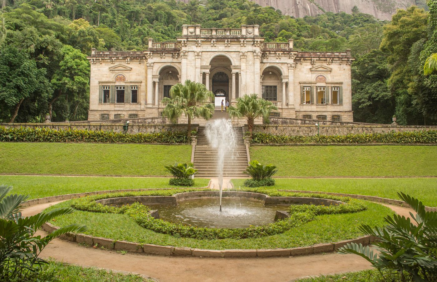 A fachada do palacete do Parque Lage sobre um gramado com chafariz redondo e palmeiras, diante da mata