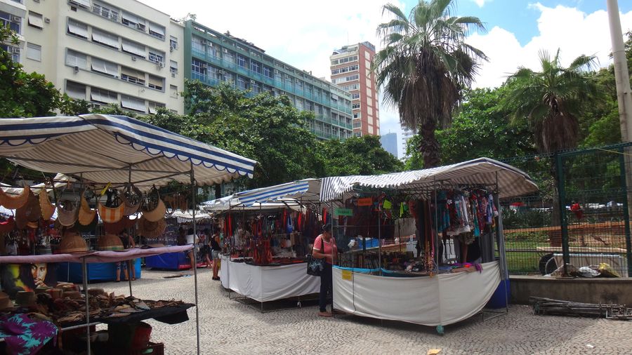 Market stalls hung with leather bags and handicrafts under palms and trees in Praça General Osório, Ipanema, with apartment blocks behind
